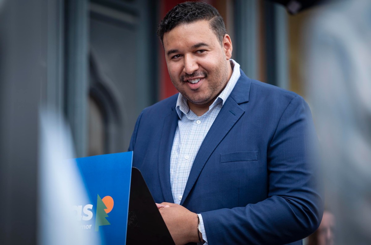 A man in a blue suit and white checkered shirt speaks at an outdoor event, smiling. He stands behind a podium with a blue sign. The background includes blurred architectural features.