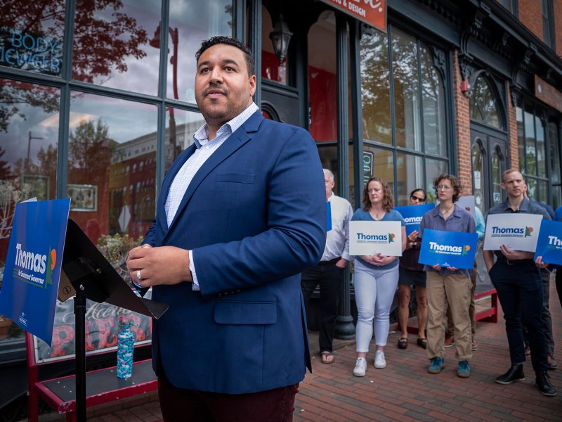 A man in a suit stands in front of a podium, addressing an outdoor gathering. Several people behind him hold signs that read "Thomas." They are in front of a building with large windows.