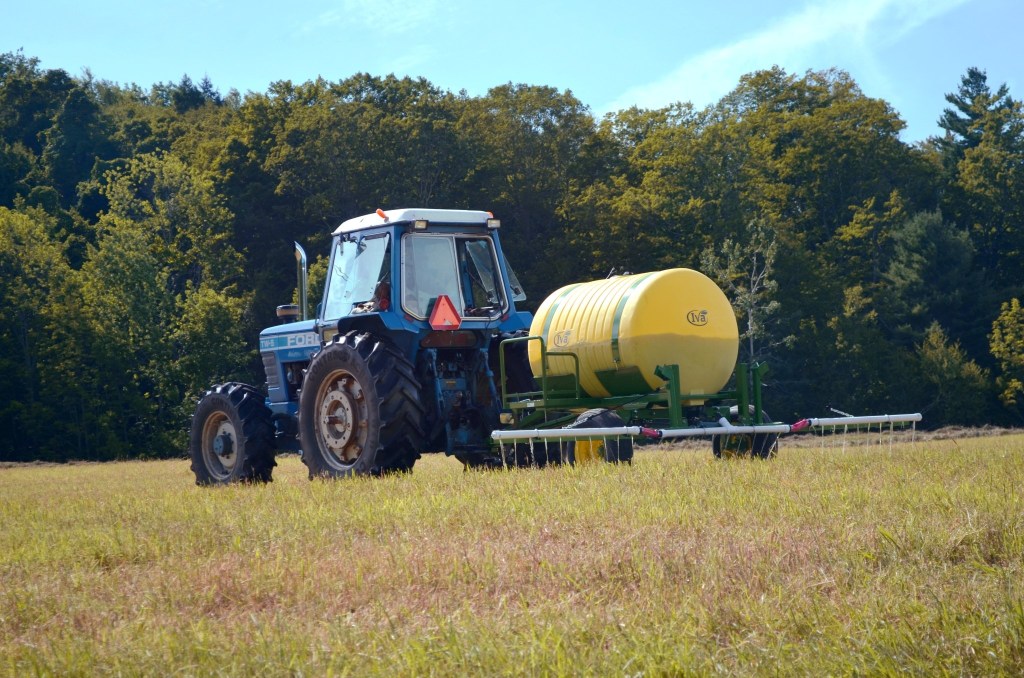 A blue tractor pulling a yellow tank sprayer across a grassy field with trees in the background under a clear sky.