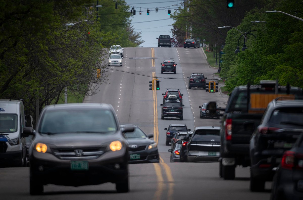 Cars driving on a tree-lined street with multiple traffic lights and lanes. Greenery is present on both sides of the road, and the sky is overcast.