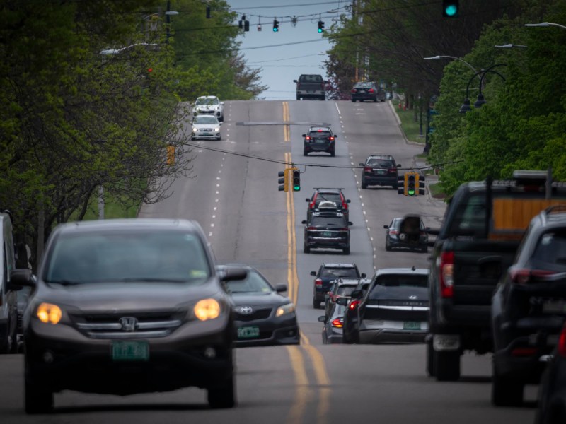 Cars driving on a tree-lined street with multiple traffic lights and lanes. Greenery is present on both sides of the road, and the sky is overcast.