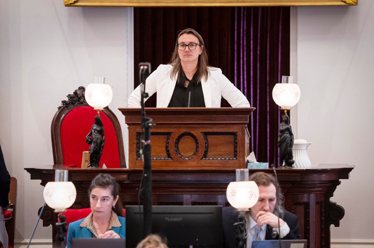 A woman stands at a wooden podium in a formal room with ornate red chairs and decorative lamps. Two other individuals are seated at desks in front of her, working on papers and computers.