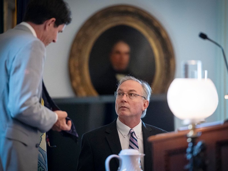 Two men in suits are engaged in conversation in a formal room. One is standing and holding a tie, and the other is seated and looking up. A portrait hangs in the background.