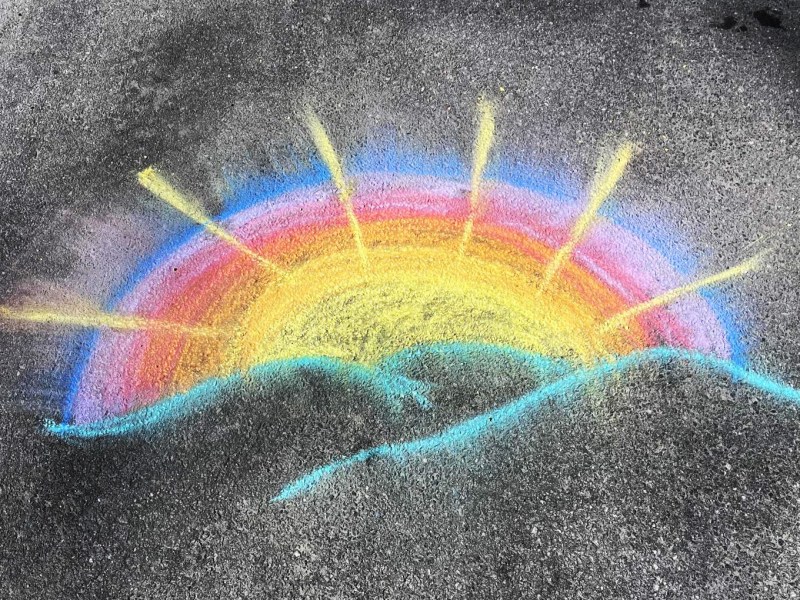 Chalk drawing of a vibrant rainbow with radiating yellow lines and a light blue wave on an asphalt surface.