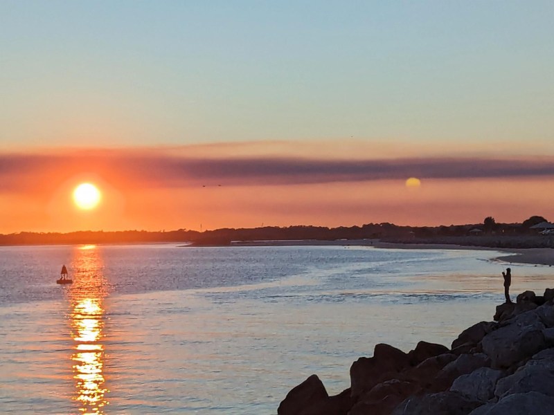 A person standing on rocks photographs the ocean at sunset, with the sun's reflection visible on the water. Another person in a small boat floats on the water near the horizon.
