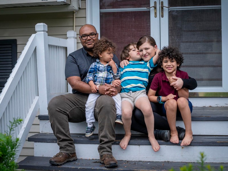 A family of five sits together on the steps outside a house. An adult man, an adult woman, and three children smile for the photo.