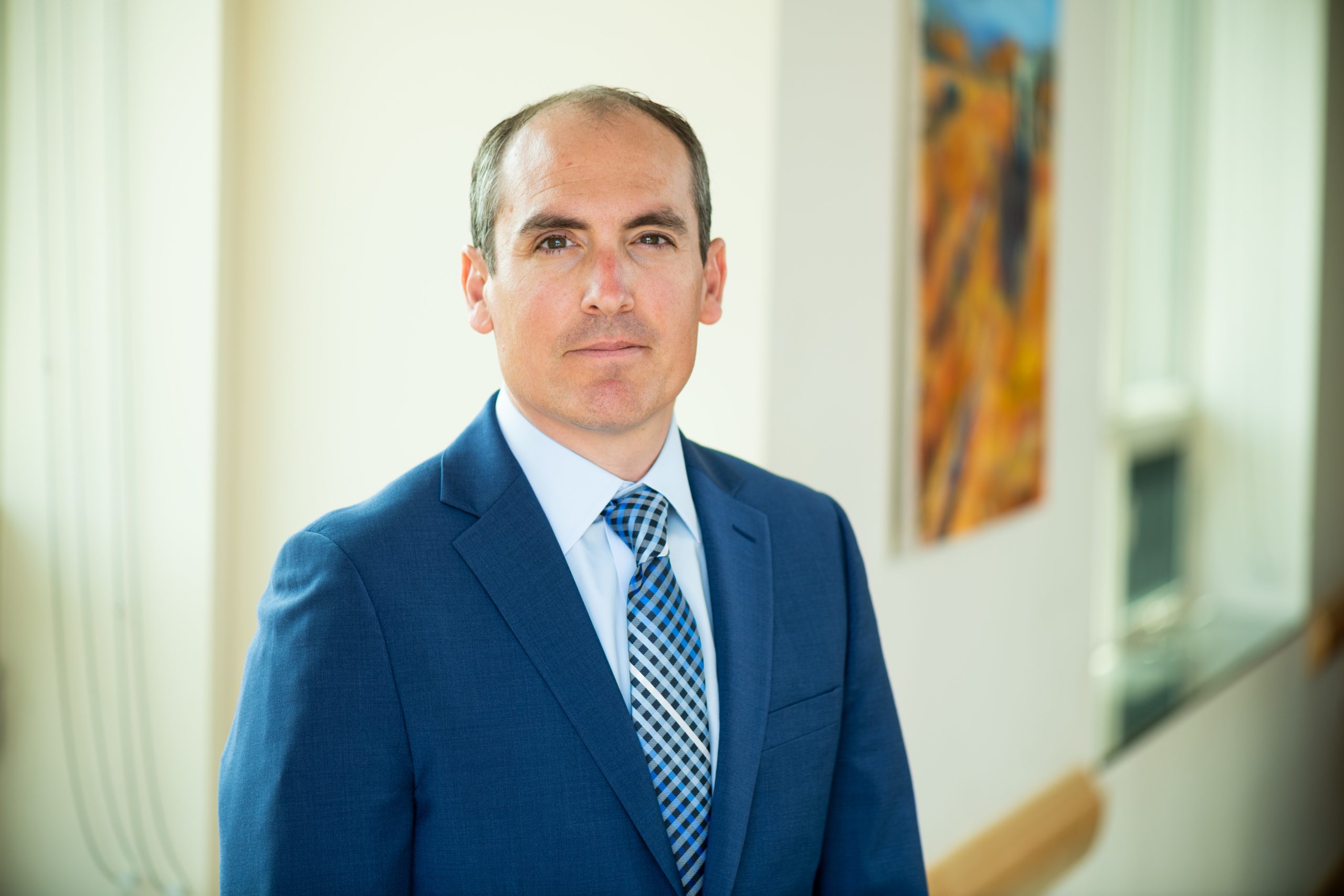 A man wearing a blue suit and tie stands indoors.