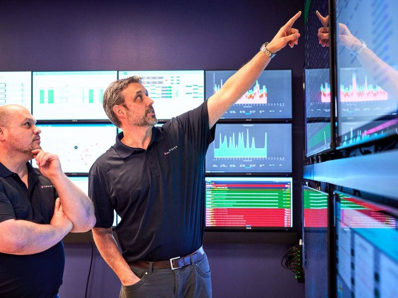two men pointing at a wall of monitors.