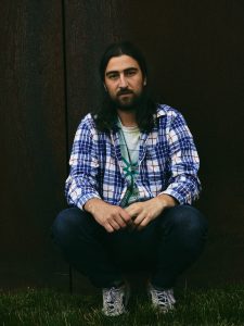 A man in a plaid shirt kneeling in front of a rusty wall.