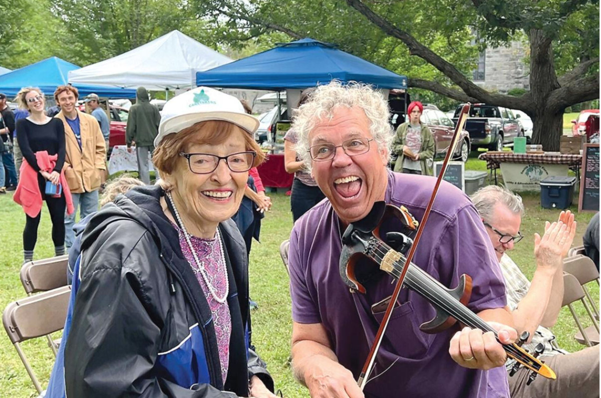A man plays the fiddle while a woman watches and another man claps in an open space.