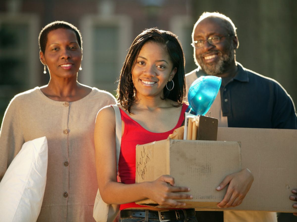 A young woman carrying a box is accompanied by two older adults, possibly her parents, outside during daylight.