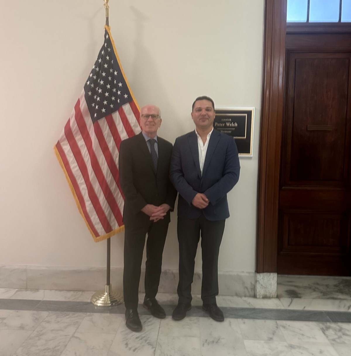 Two men standing in front of an office door with a nameplate. An American flag is on the left side of the image. They are in a hallway with marble flooring.