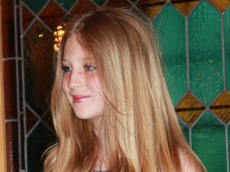 A young person with long, straight blonde hair stands in front of a stained glass window featuring green and amber geometric patterns.