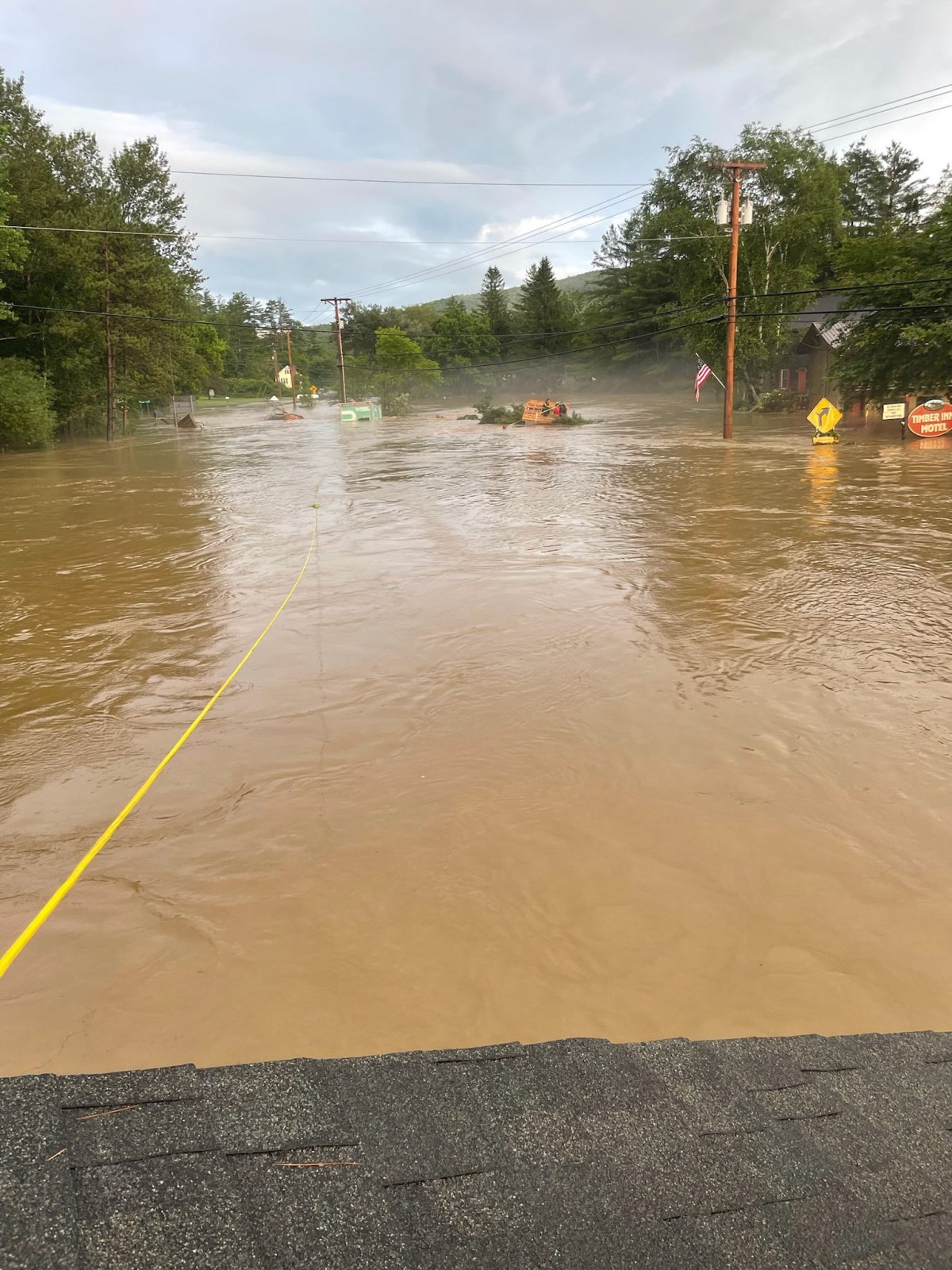 A flooded street with muddy water covering the road and surrounding area. Trees and utility poles are partially submerged. Yellow ropes are stretched across the scene.