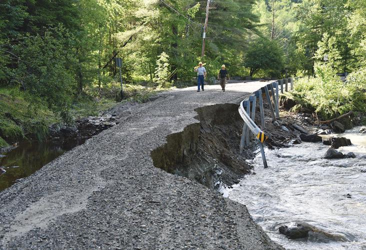 Two individuals walk along a partially eroded gravel road beside a stream, with a damaged guardrail and dense forest in the background.