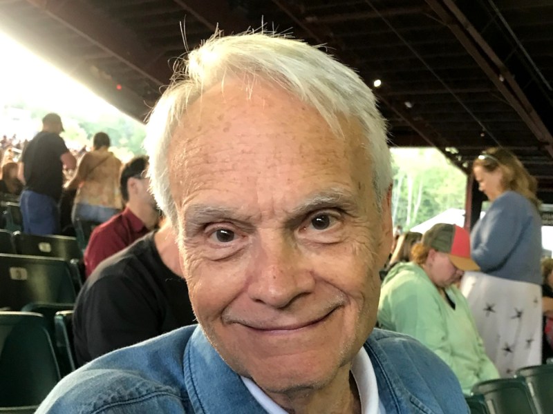 An elderly man with white hair and a denim jacket smiles at the camera while seated in an outdoor venue. Several other people are visible in the background.
