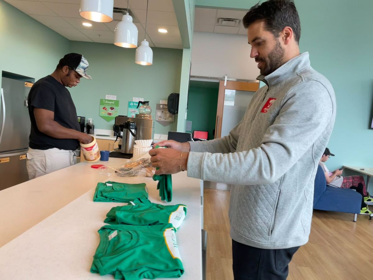 Two men standing at a counter: one scooping ice cream into a cup, and the other folding green shirts. The room has teal walls, pendant lights, and a couch in the background.