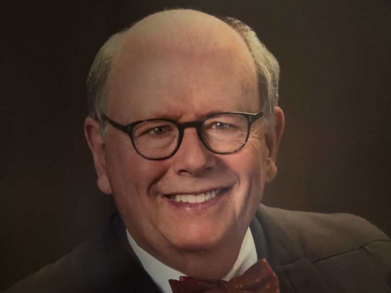 A smiling older man with glasses and thinning hair, dressed in a suit with a bow tie, is shown against a dark background.