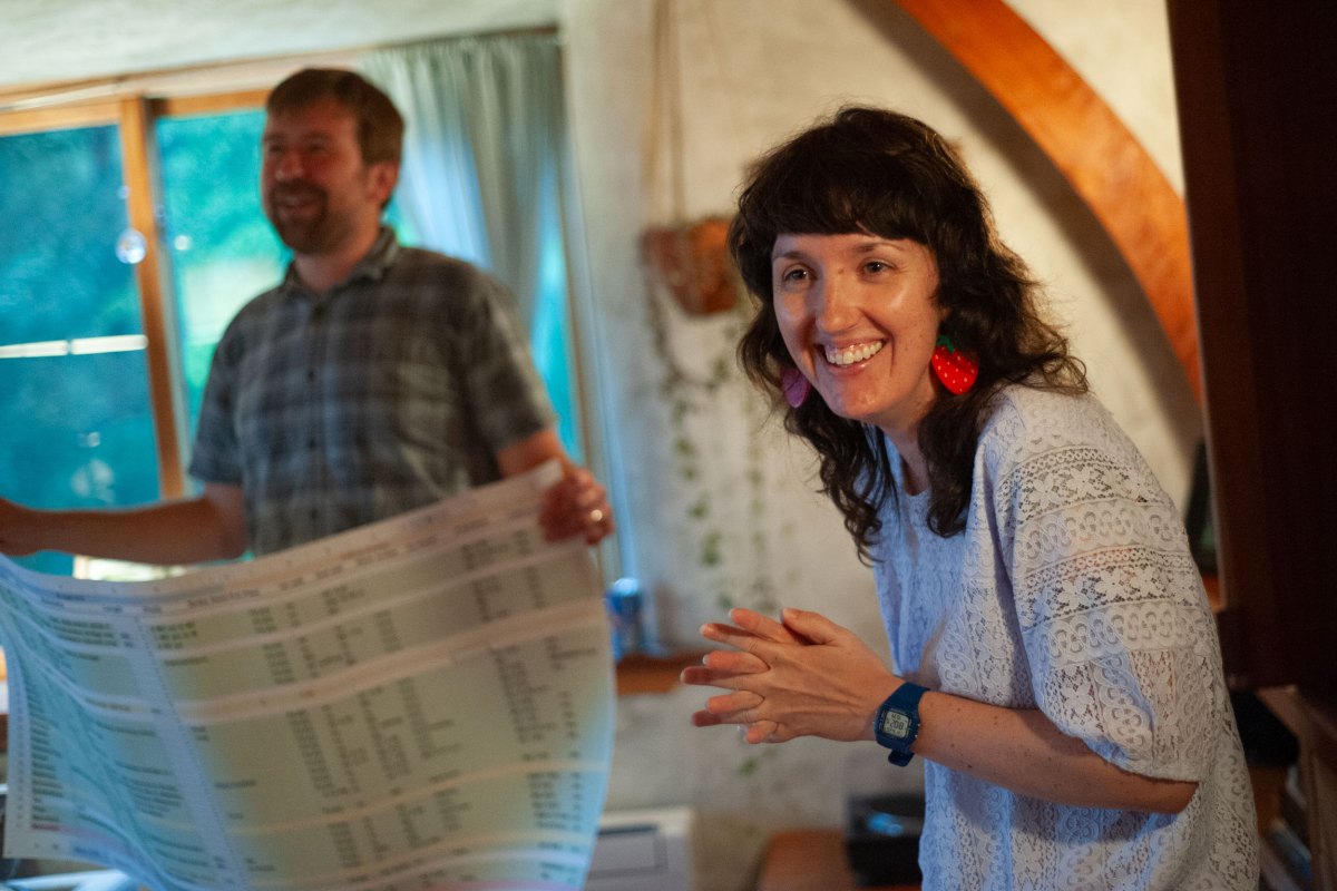 A woman with a white shirt and strawberry earrings stands on the right side of a man in a plaid shirt holding up a giant spreadsheet.