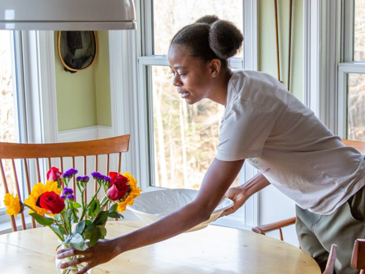 A person sets a table with a plate near a vase of colorful flowers in a room with large windows.