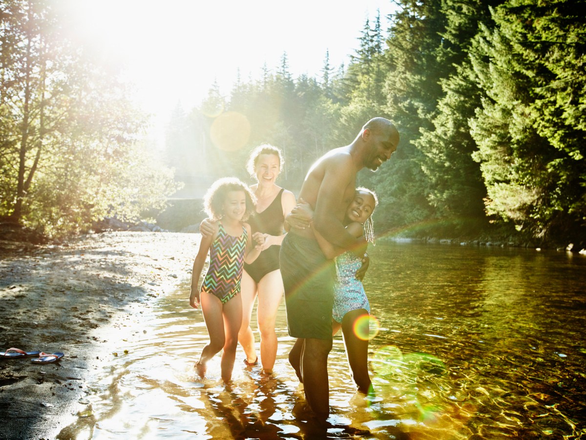 A family of four, two adults and two children, standing and playing in a shallow river surrounded by trees, illuminated by sunlight.