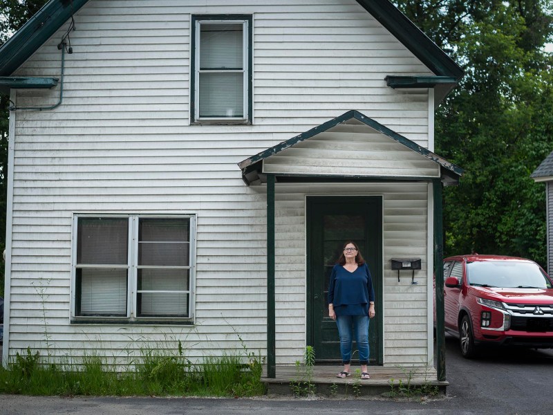 A woman stands on the front steps of a small white house with a double-pane window on the left. A red SUV is parked to the right of the house.