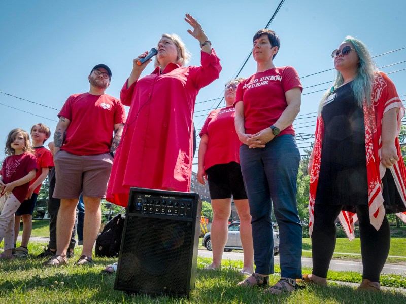 A person in a red dress speaks into a microphone while standing next to several people wearing red shirts on a sunny day. They are outdoors with a speaker in front of them.