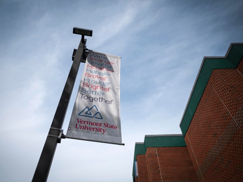 A flagpole in front of a brick building displays a banner reading "We're Stronger, Healthier, Happier, Braver, Brighter, Together." The banner also includes the Vermont State University logo.