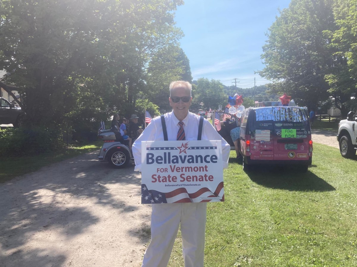 A person stands outdoors holding a "Bellavance for Vermont State Senate" sign. Several decorated vehicles with balloons and an inflatable unicorn are visible in the background.