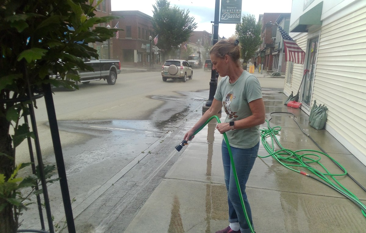 A woman in a green shirt and jeans uses a hose to wash the sidewalk on a street with parked cars and buildings in the background.