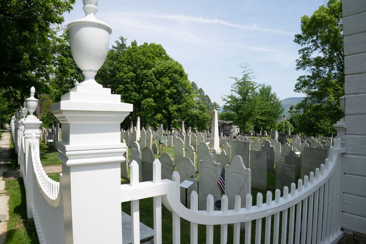 White picket fence surrounding a cemetery with rows of gravestones, green trees, and hills in the background.