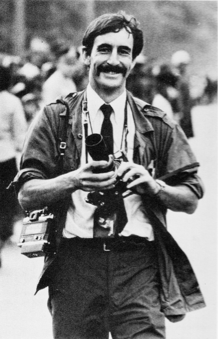 Black and white photo of a smiling man with a mustache holding a camera lens and wearing multiple cameras around his neck. He is walking outdoors with people in the blurred background.