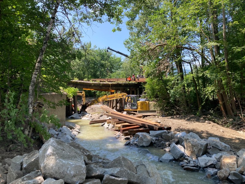 Construction workers and machinery are at a bridge rebuilding site in a wooded area, with heavy equipment and materials visible around a streambed.