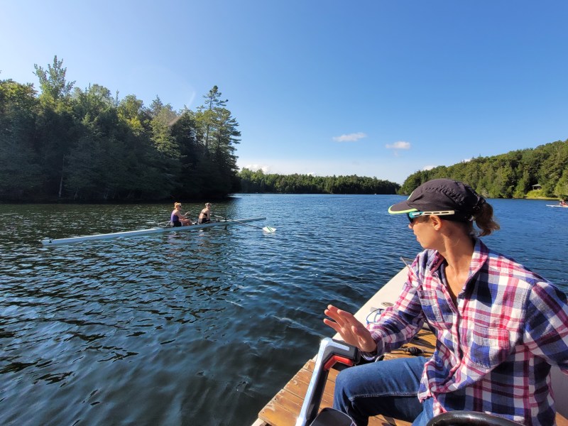 Person in a plaid shirt and cap on a boat looking at two rowers on a calm lake surrounded by forest under a clear blue sky.