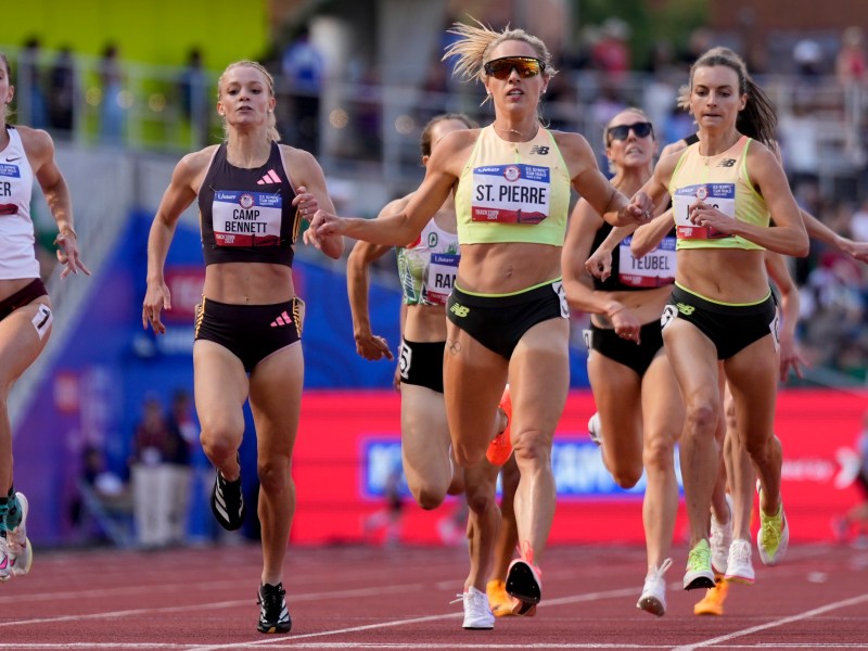 A group of athletes sprinting toward the finish line on a track during a race. One runner in the center, wearing a yellow top, appears to be in the lead. Others are close behind.
