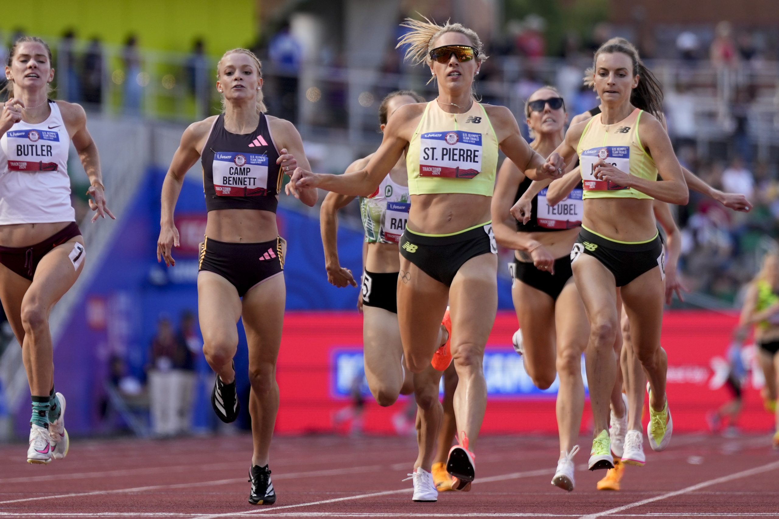 A group of athletes sprinting toward the finish line on a track during a race. One runner in the center, wearing a yellow top, appears to be in the lead. Others are close behind.
