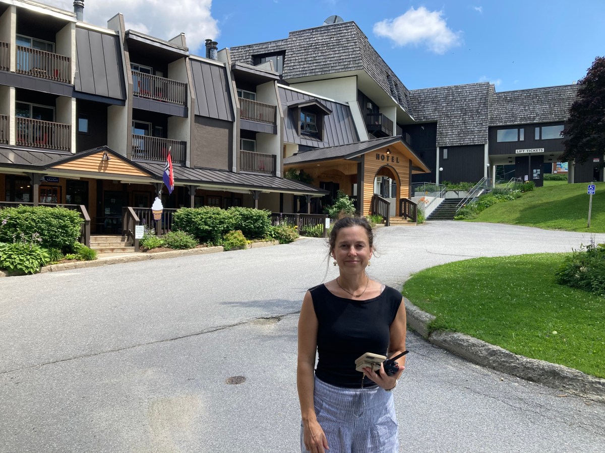 A woman in a black top and light-colored pants stands on a paved path in front of a hotel building with multiple balconies, surrounded by greenery. She holds a book and her phone in her hands.