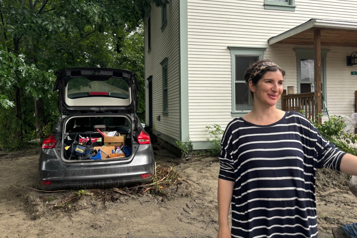 A woman stands outside near a white house, with a car parked beside it. The car's trunk is open, revealing various tools and boxes inside. The area appears muddy and wet, suggesting recent flooding.
