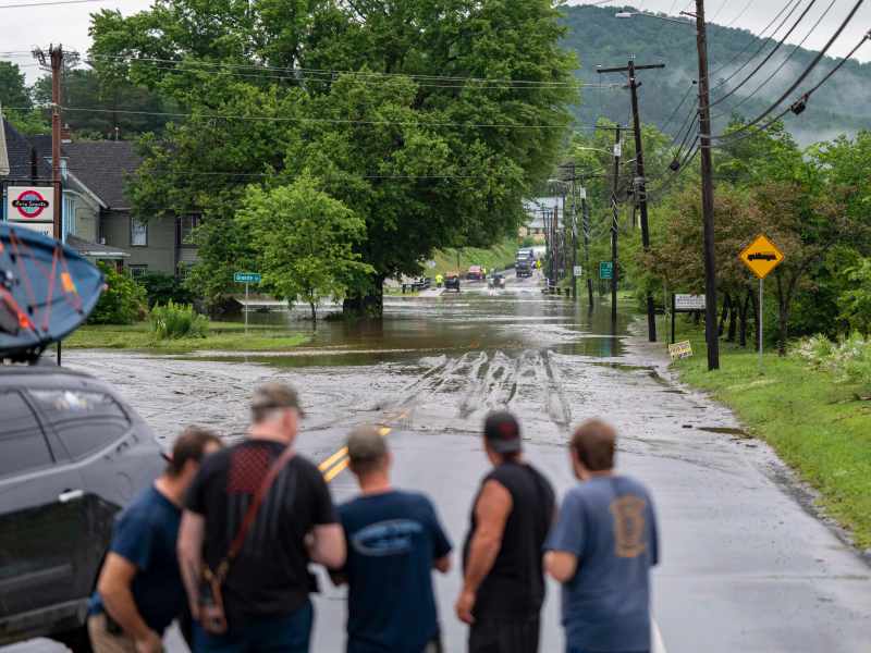 A group of people stands near a flooded street, observing the water covering the roadway. Several vehicles and houses are in the background, with overcast skies and greenery surrounding the area.