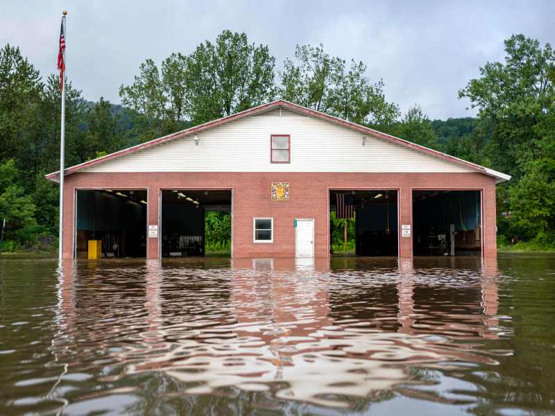 A brick building with three garage doors is partially submerged in floodwater, with trees and a flagpole in the background.
