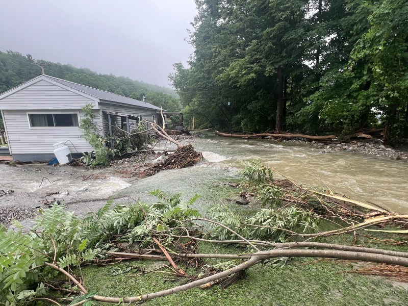 A house is partially submerged in floodwater, surrounded by debris and fallen branches, next to a rapidly flowing creek in a forested area.