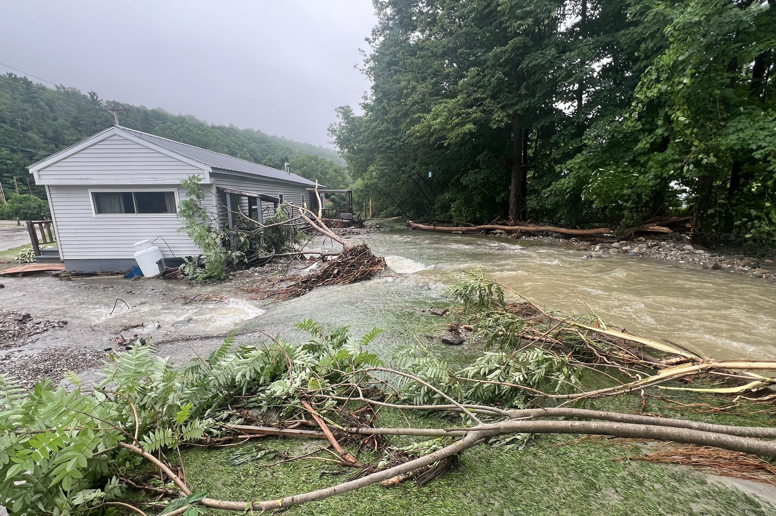 A house is partially submerged in floodwater, surrounded by debris and fallen branches, next to a rapidly flowing creek in a forested area.