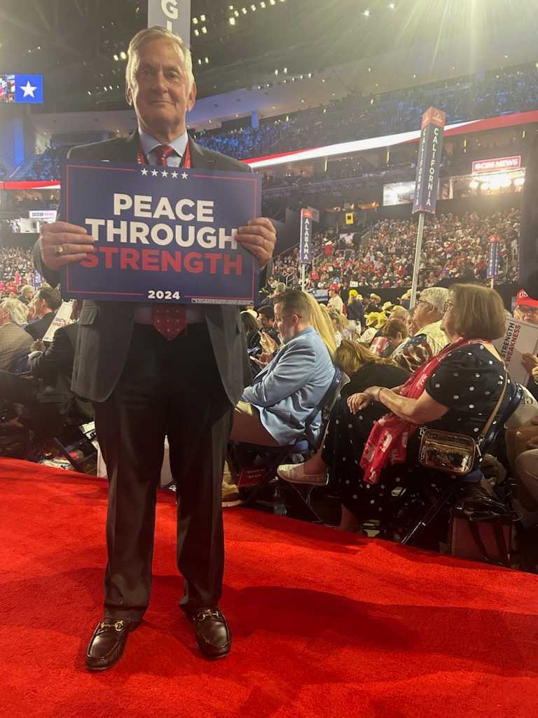 A man in a suit stands on a red carpet holding a sign that reads "Peace Through Strength 2024" at a crowded indoor event.