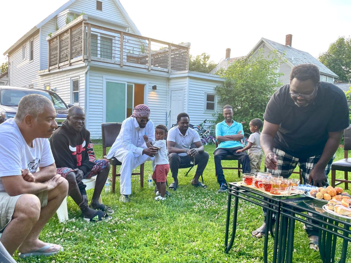 A group of people sit and stand around a table with food and drinks in a backyard, engaging in conversation and activities.