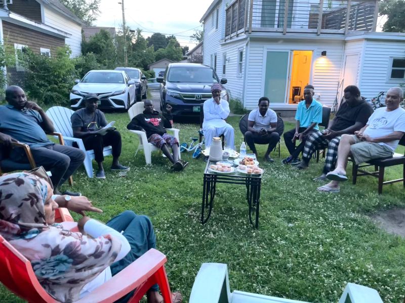 A group of people sit in a circle of chairs outside in a backyard, engaging in conversation. Food and drinks are placed on a small table in the center.