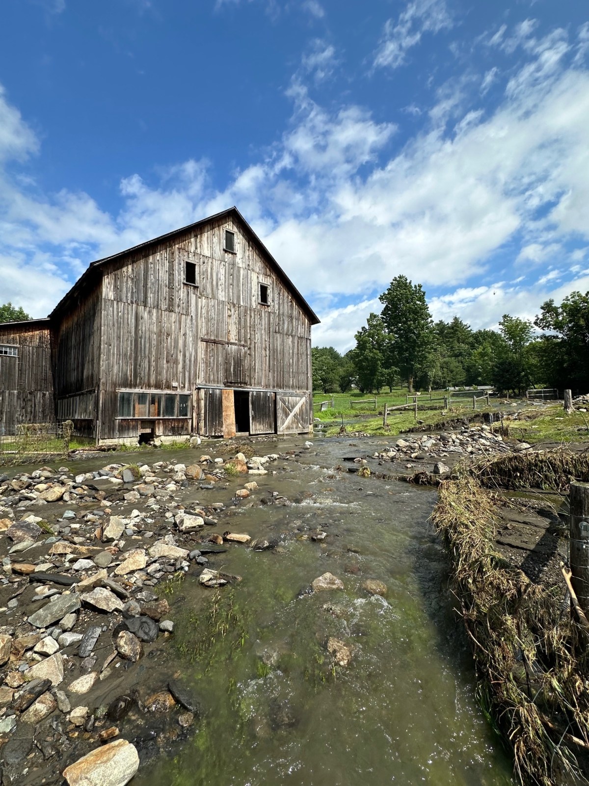 A weathered wooden barn stands beside a rocky stream under a partly cloudy sky, with green trees and grass in the background.