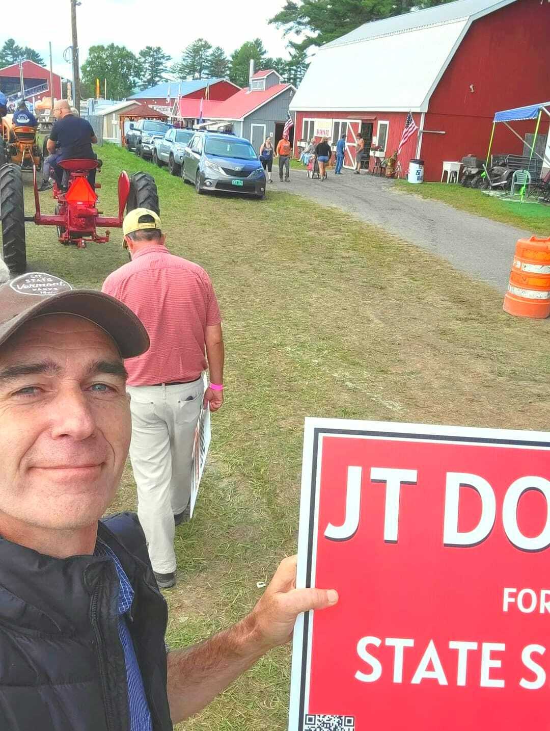 A person holds a campaign sign reading "JT Dodd for State Senate" while standing in a line. People and a tractor are in view, with red buildings and tents in the background.