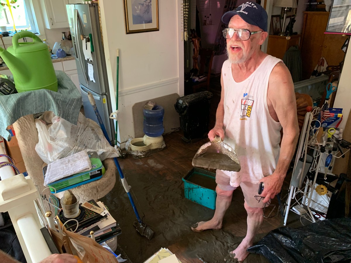 An elderly man wearing a cap and glasses, stands in a cluttered room with a flooded floor, holding a scoop shovel and a small flashlight. Various household items are scattered around.