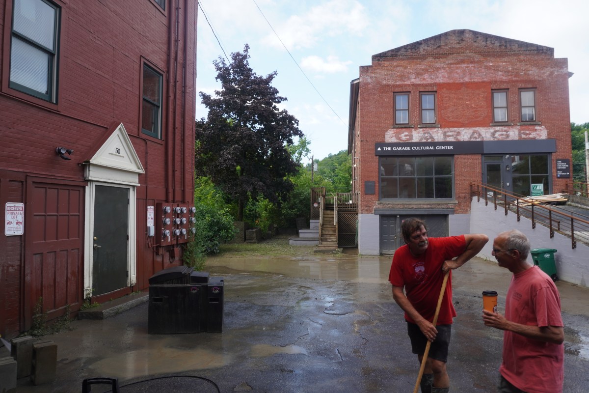 Two people in red shirts stand in an alley with wet ground between brick buildings, one holding a broom and wiping his brow, and the other holding a drink.