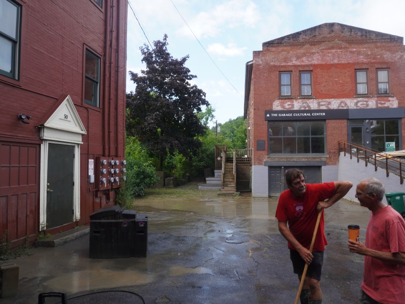 Two people in red shirts stand in an alley with wet ground between brick buildings, one holding a broom and wiping his brow, and the other holding a drink.